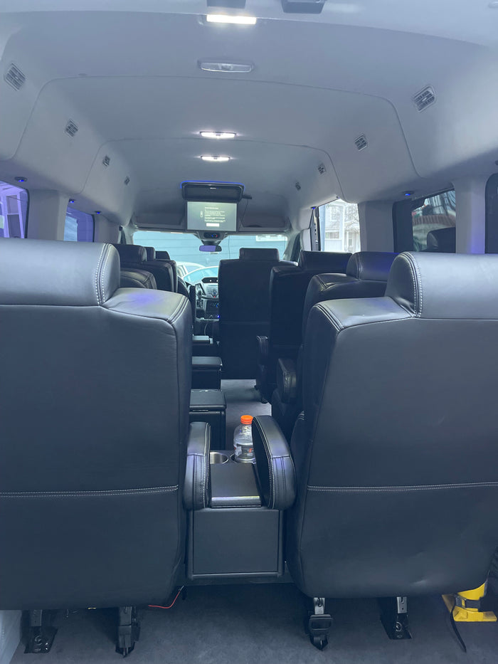 Comfortable interior of a passenger van, ideal for RV rental. Features plush black leather seats, overhead screen, and ample lighting.