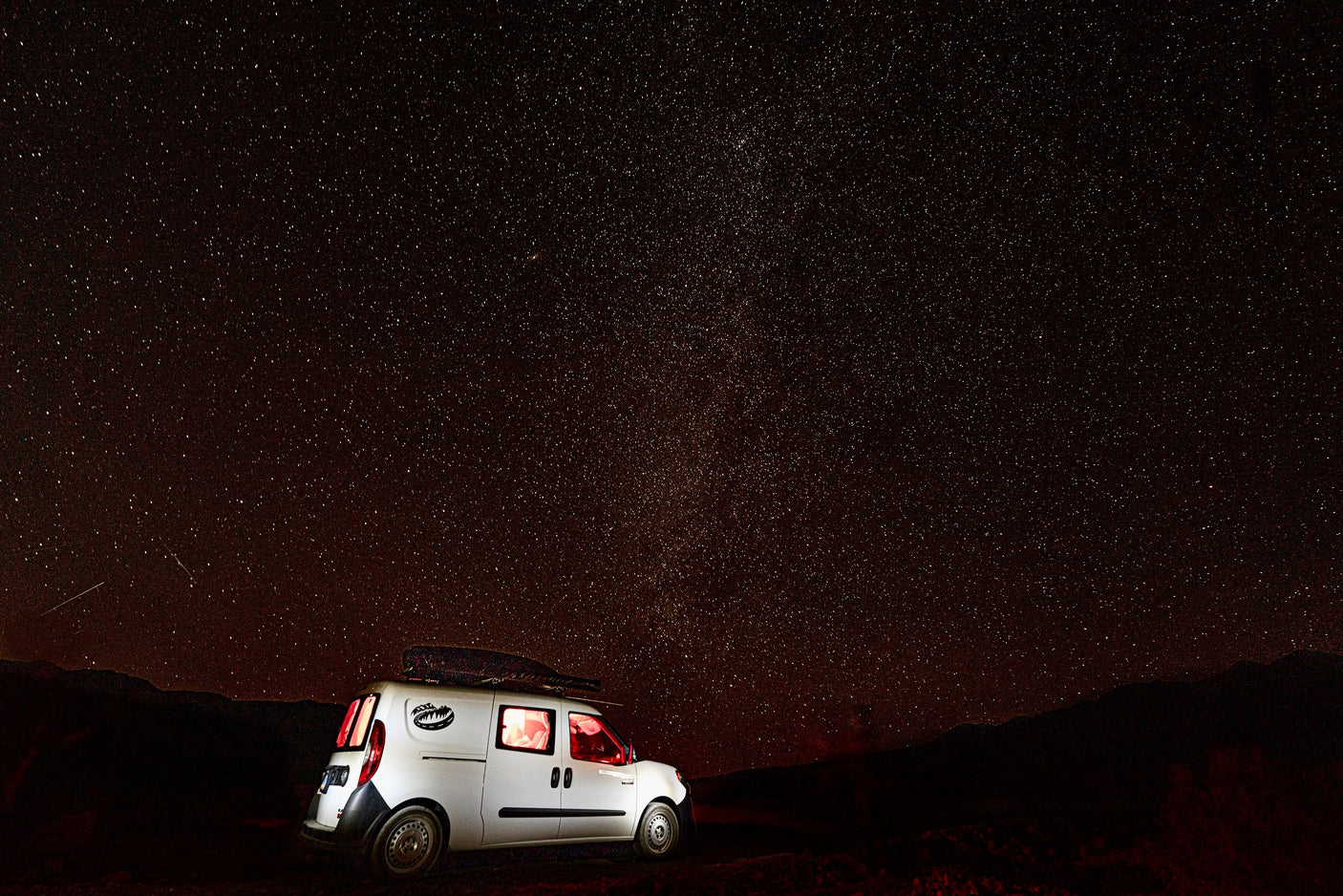 White RV rental camper van with glowing interior lights under a brilliant starry night sky with distant mountains.