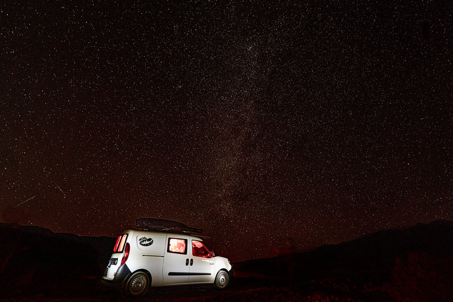 White RV rental camper van with glowing interior lights under a brilliant starry night sky with distant mountains.