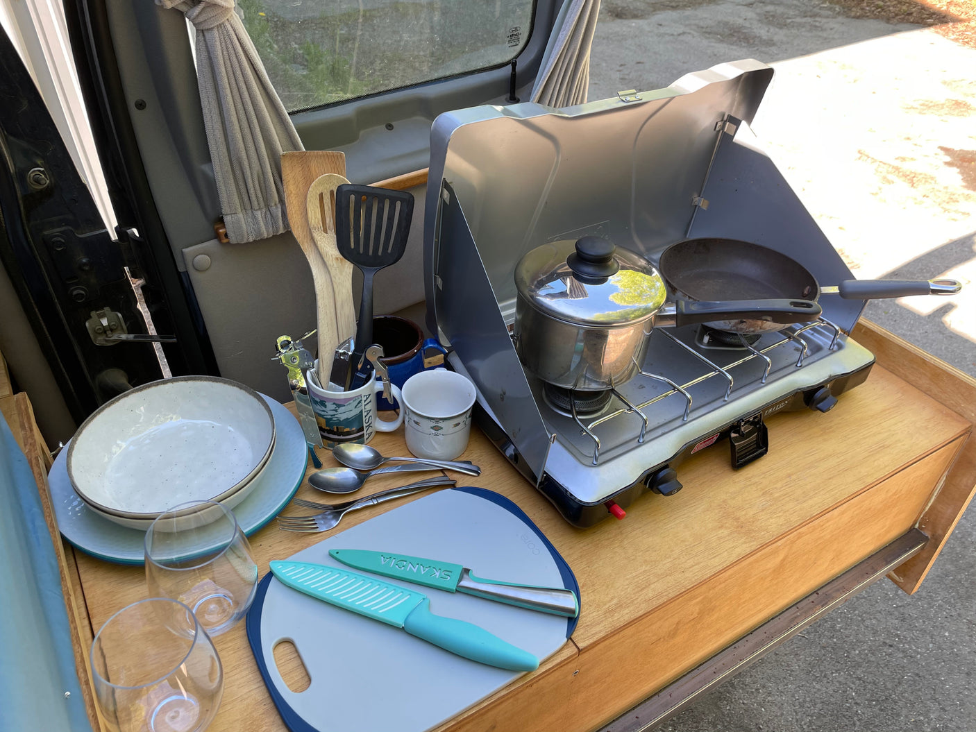 RV rental: A compact, pull-out camp kitchen with a Coleman stove, pots, pans, plates, and utensils, ready for outdoor cooking.