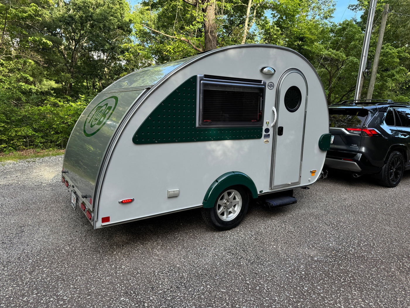 A white & green teardrop RV rental, perfect for outdoor travel, is hitched to an SUV on a gravel path surrounded by trees. — photo 5