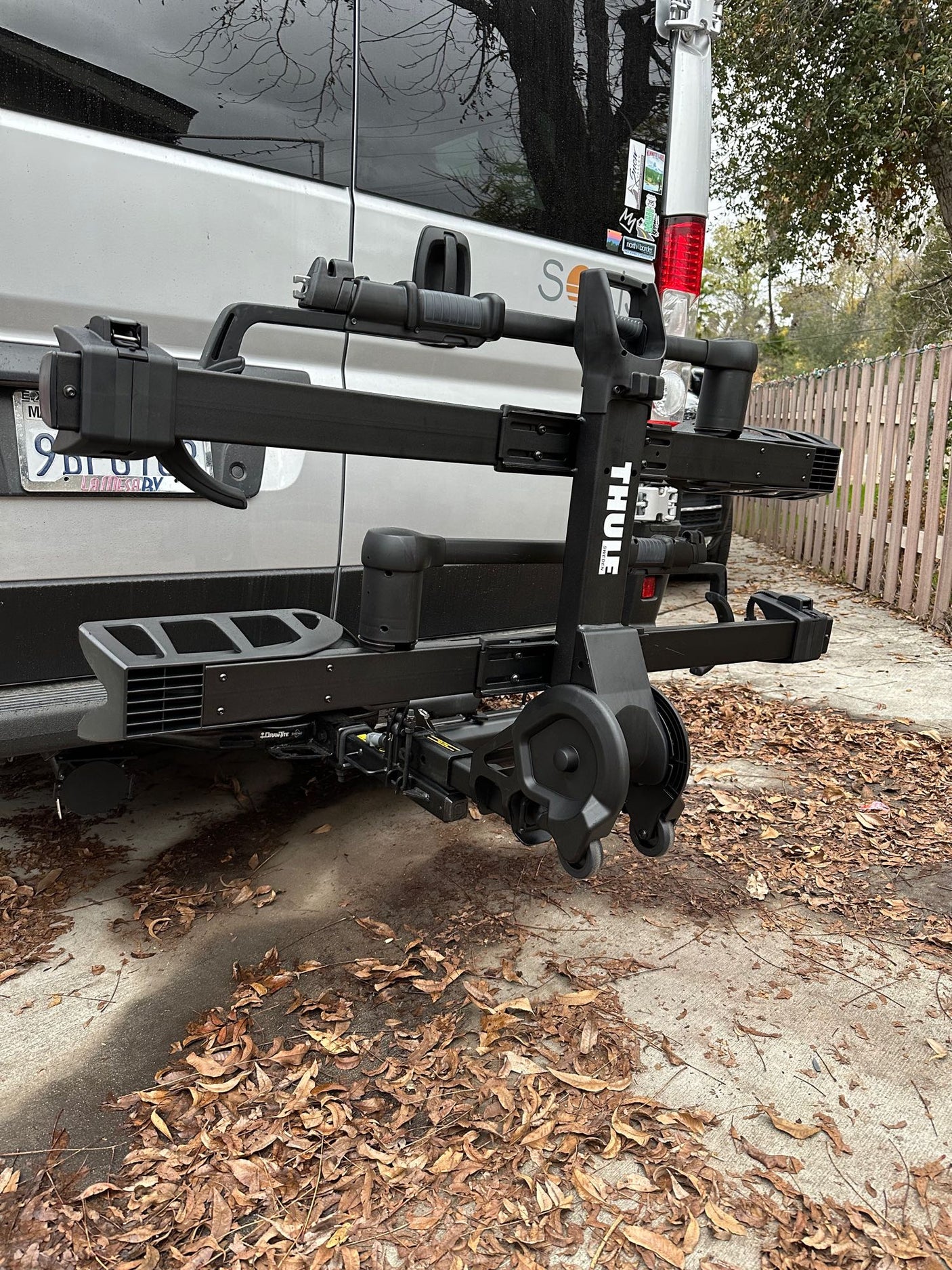 Rear view of a gray RV rental, featuring a folded Thule bike rack. Leaves cover the concrete ground.