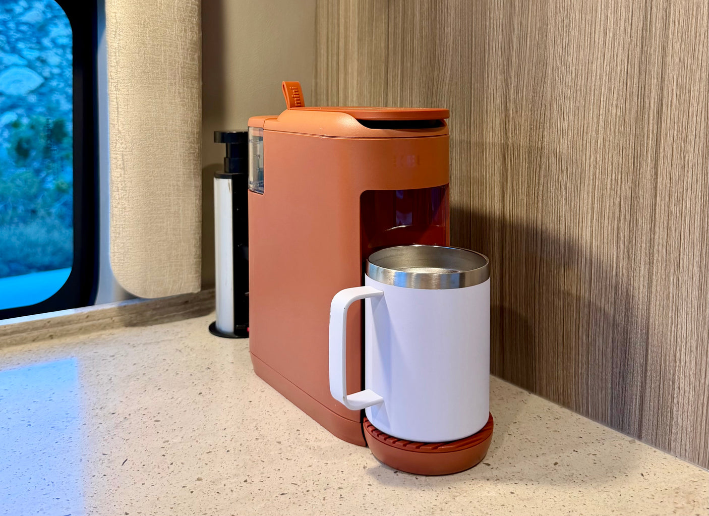 Convenient coffee setup in an RV rental: a compact coral coffee maker and white mug on a speckled counter by a window.