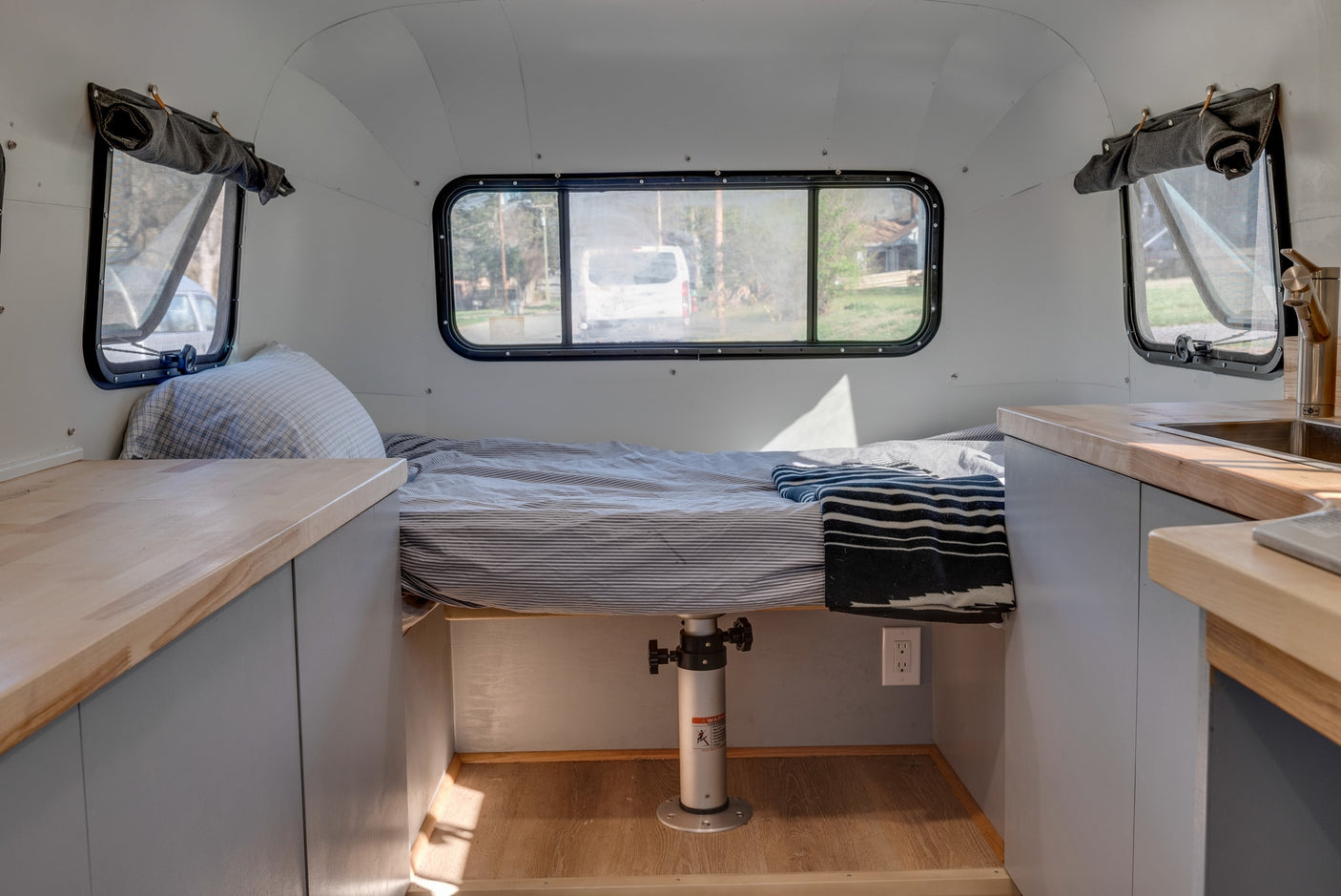 An RV rental's interior shows a striped bed, wooden counters with a sink, and windows, offering a cozy, functional space.