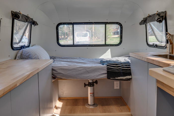 An RV rental's interior shows a striped bed, wooden counters with a sink, and windows, offering a cozy, functional space.