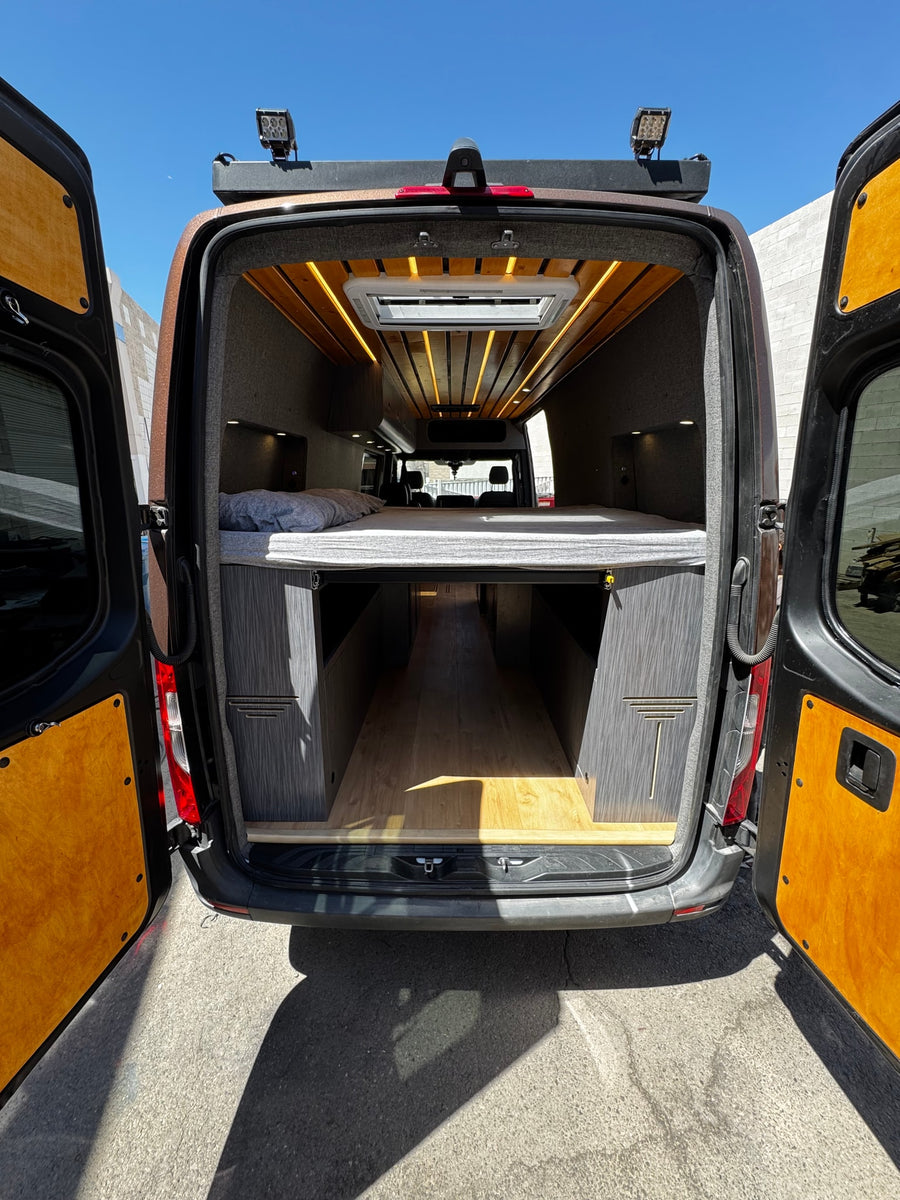 Modern van conversion RV rental: rear view revealing a comfortable bed, under-bed storage, and wood-slat ceiling with lights.