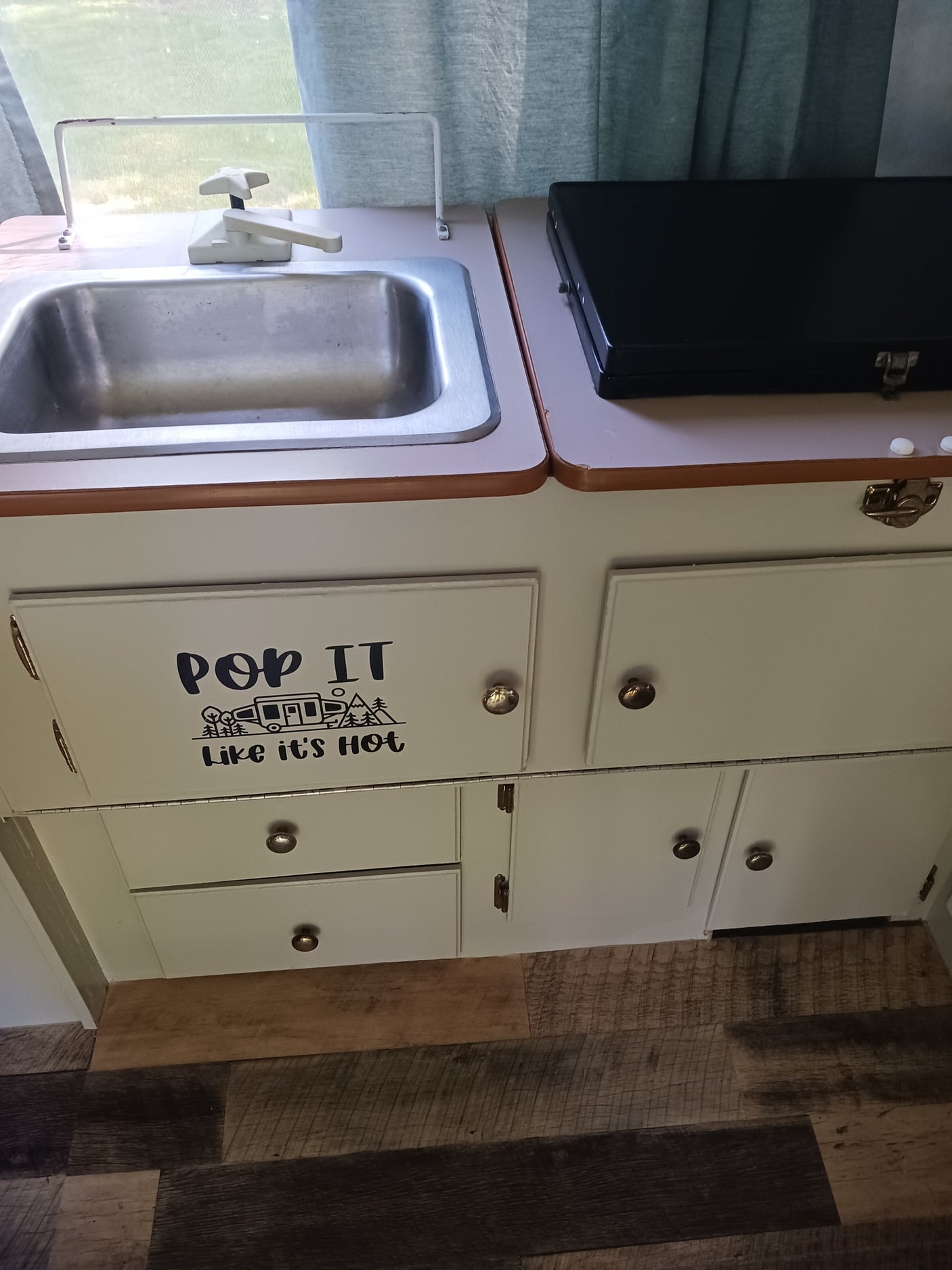 RV rental kitchen: compact area with a stainless steel sink, black portable stove, and cabinets, one with a "Pop It Like It's Hot" camper decal.