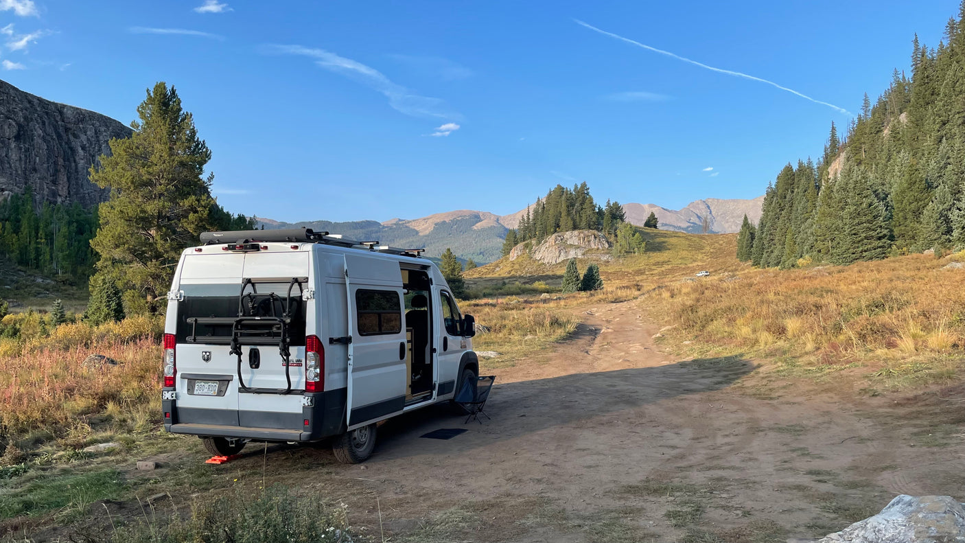 White RV rental van parked on a dirt road in a scenic mountain landscape. Trees, dry grass, and peaks under blue sky.
