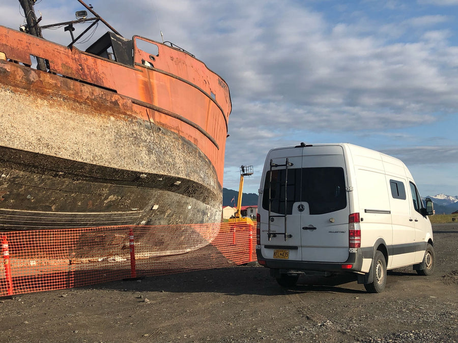 rusty ship in dry dock under a cloudy sky. Unique Alaskan scene.|Adventure awaits! An RV rental Sprinter van parked on a rocky riverbed at sunset