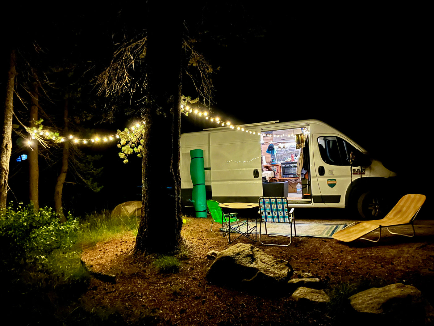 Night view of an RV rental campsite: bright van interior, string lights, and outdoor chairs amidst trees, creating a cozy scene.