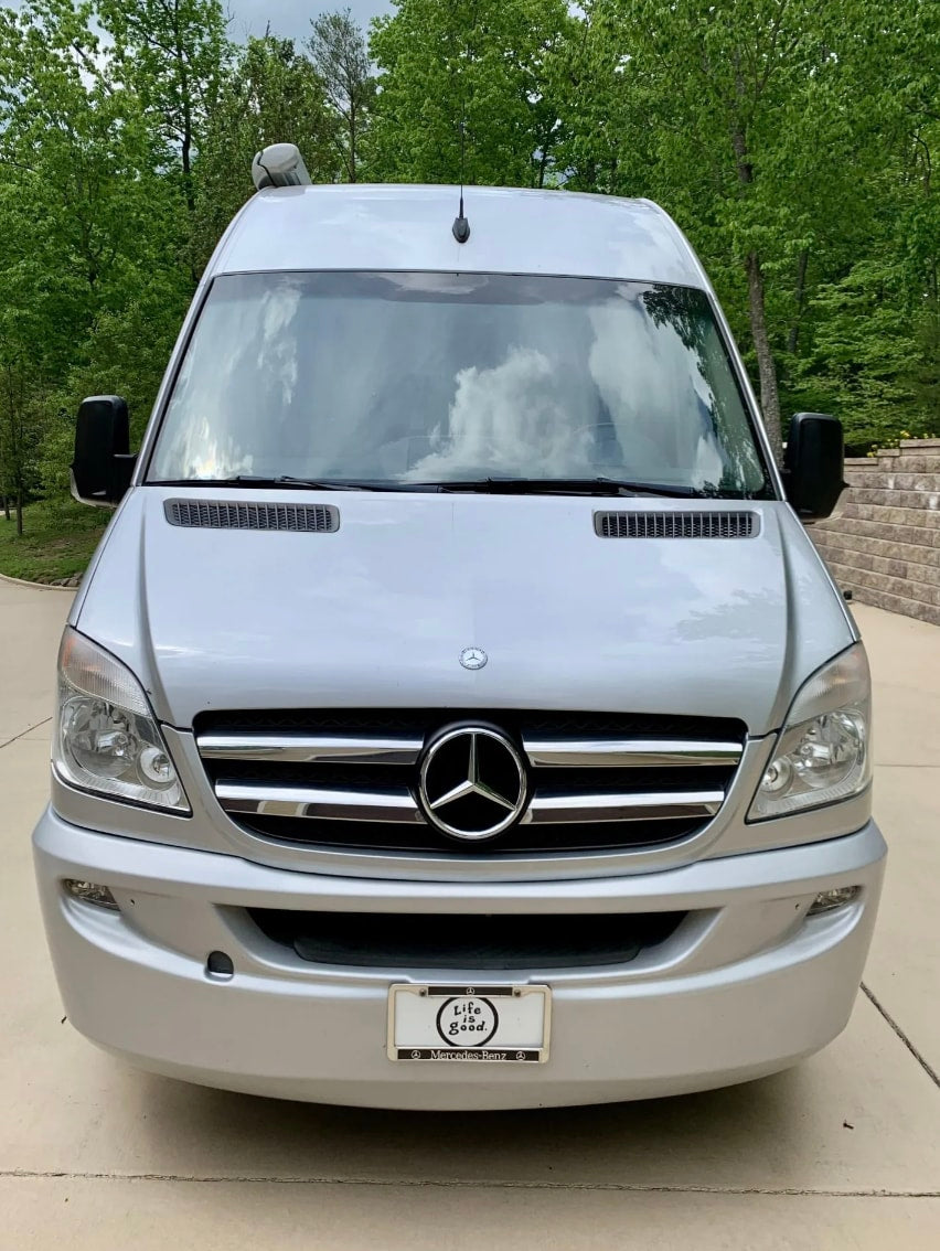Front view of a silver Mercedes-Benz Sprinter RV rental. This luxury travel van is parked against a backdrop of green trees.