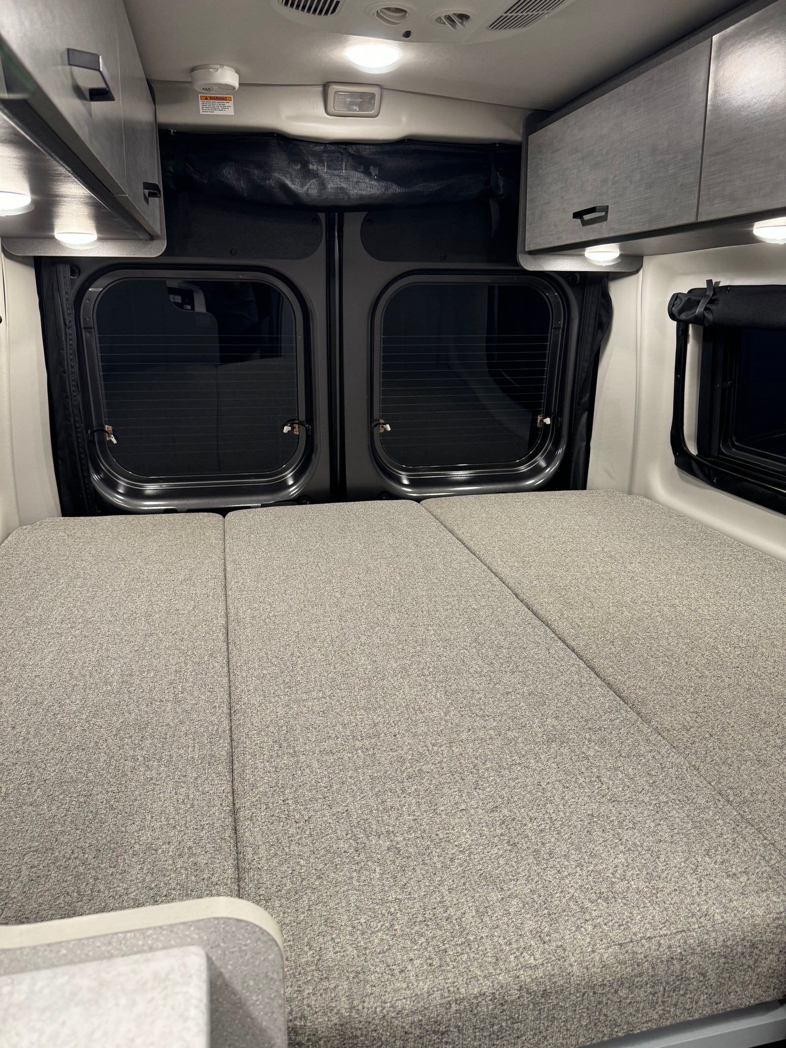 Comfortable RV rental interior featuring a spacious bed, overhead storage cabinets, and rear windows, ready for your trip.
