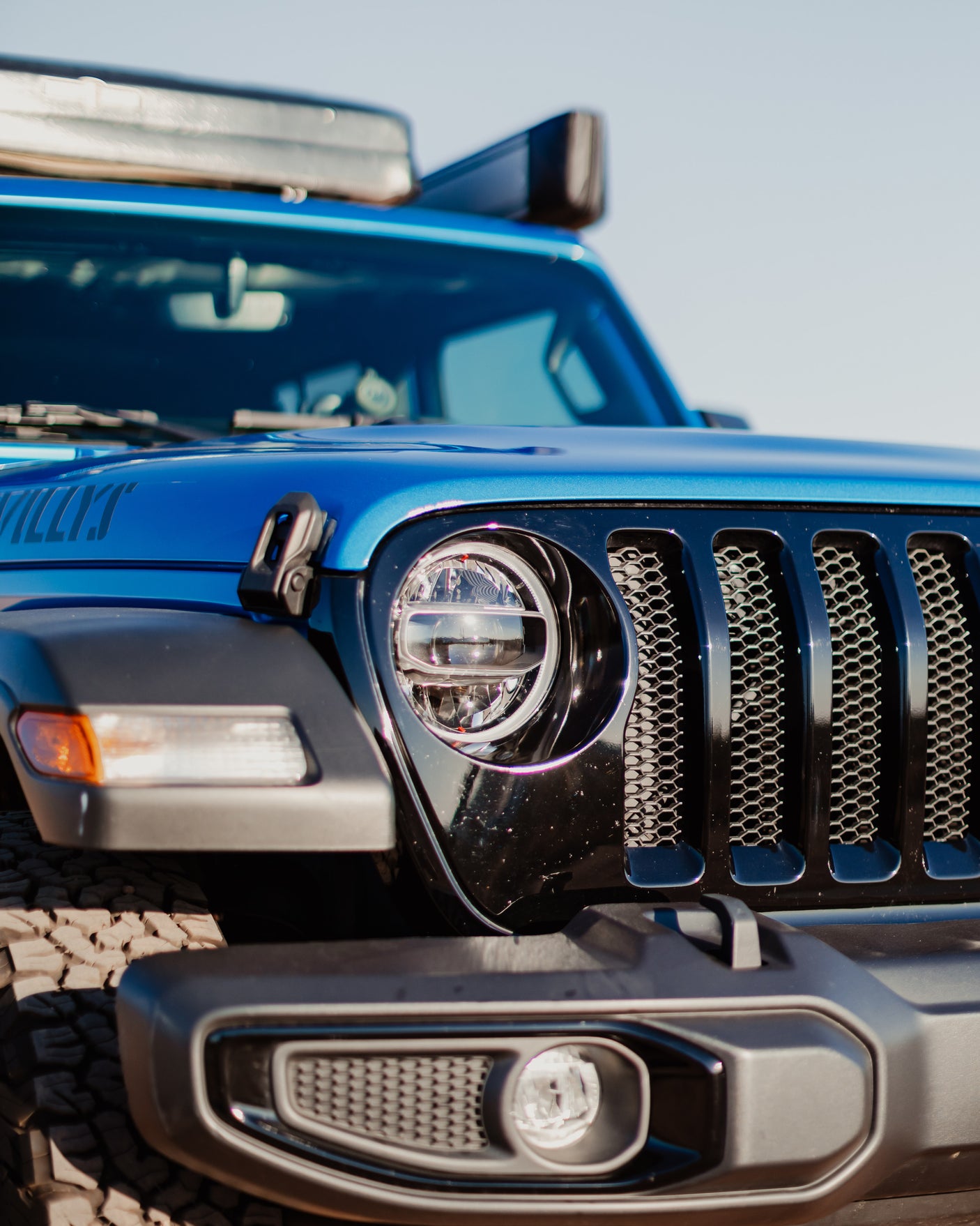 Close-up of a blue Jeep Wrangler Willys' front, showing headlight, grill, and roof storage. Explore with an rv rental.