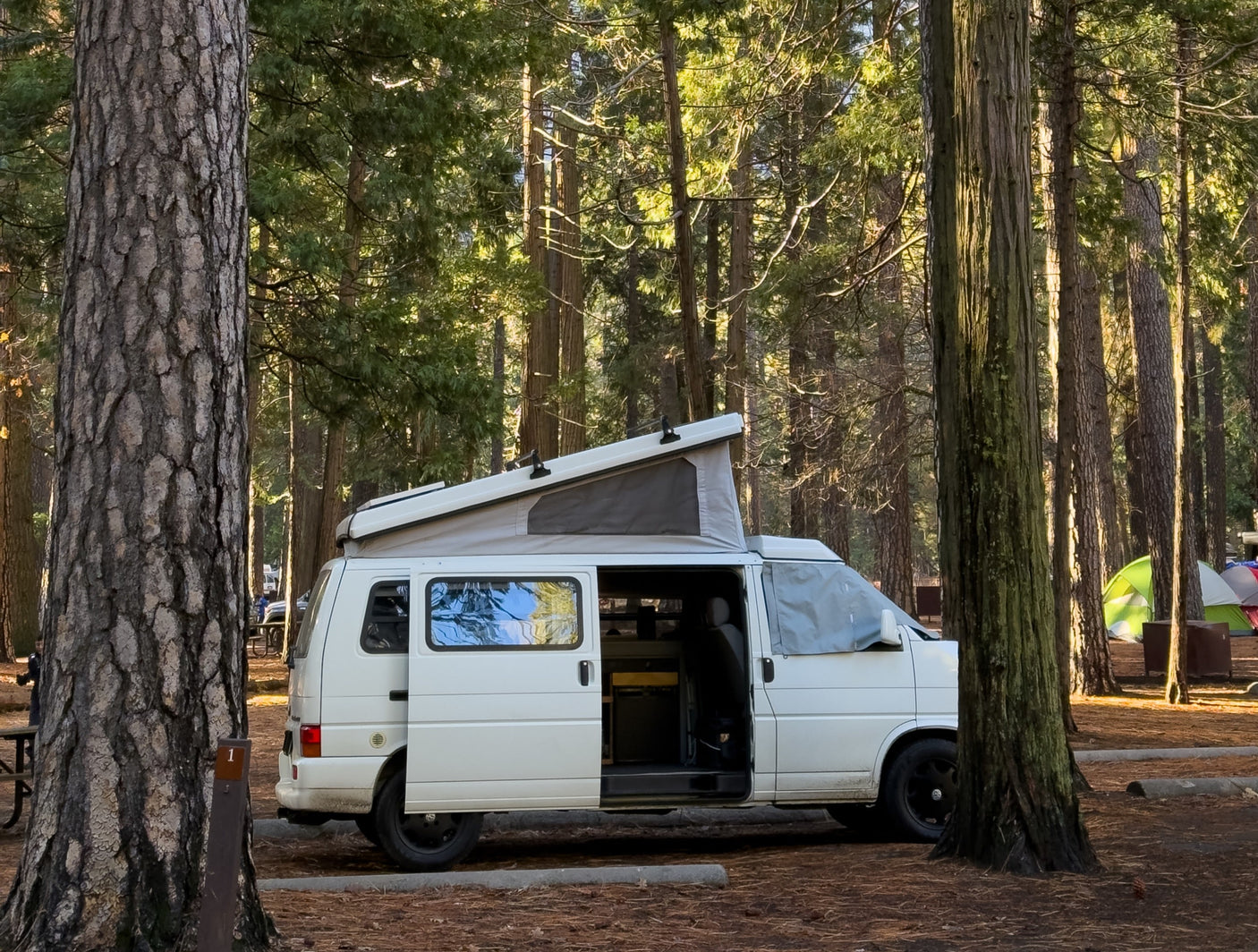 An RV rental pop-top camper van at a sunny forest campsite with its side door open, surrounded by tall trees and tents.