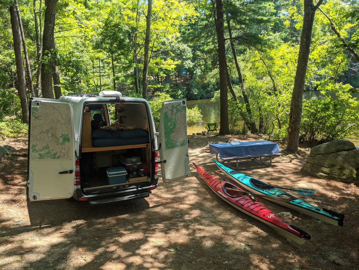 An RV rental camper van, doors open to reveal bed & storage, sits with kayaks by a lake, ready for campsite adventure.