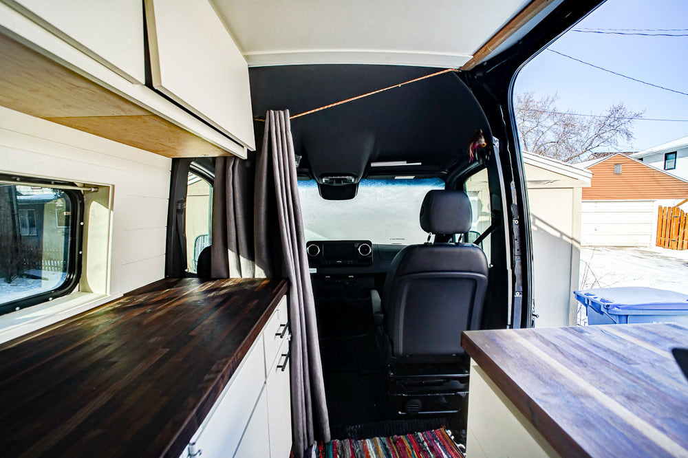 Inside this modern custom camper van, perfect for your next rv rental. Features wood counters, white cabinets, and driver's cabin view.