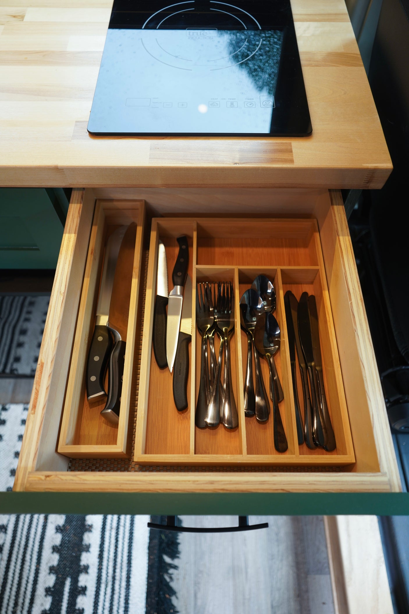 Top-down view of an RV rental kitchen: wood countertop, induction cooktop, and neatly organized cutlery drawer.