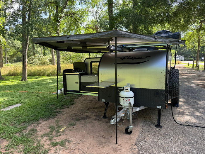 An RV rental, an off-road camper with an extended awning and outdoor kitchen, is set up at a leafy campsite.