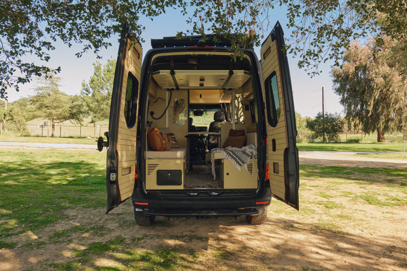Black camper van rv rental with open rear doors showing a cozy, wood-paneled interior. Parked in a grassy outdoor setting.