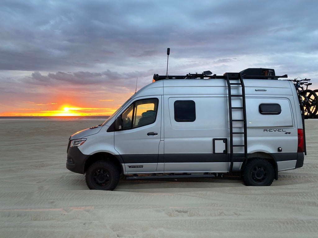 Experience van life freedom! A silver RV rental, a Winnebago Revel 4x4, parked on a beach at a vibrant sunset.