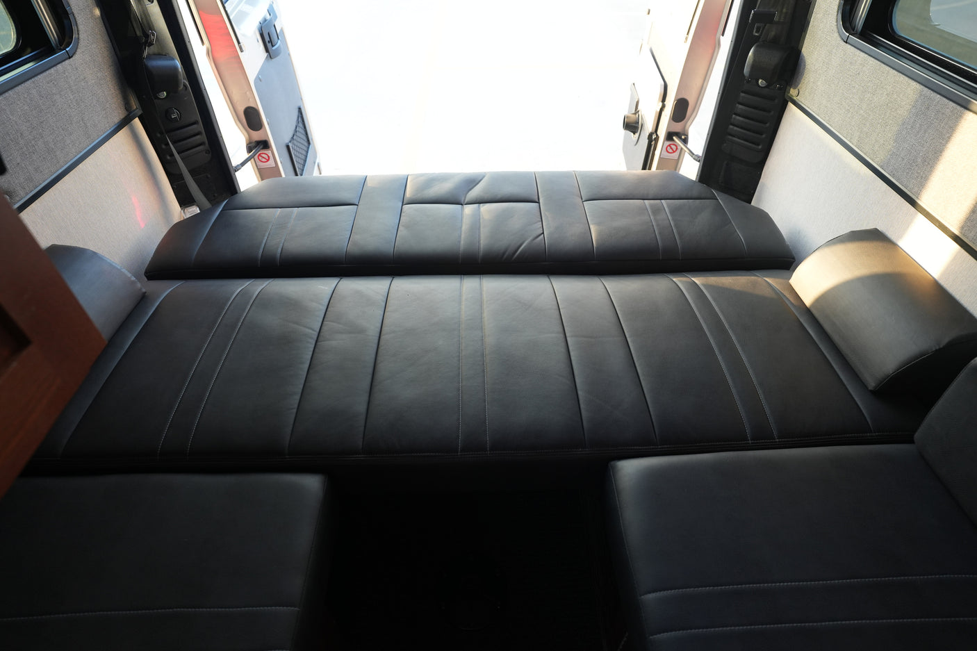 Spacious RV rental interior: black leather seating converts to a bed. Back doors open, bright light fills the camper van.