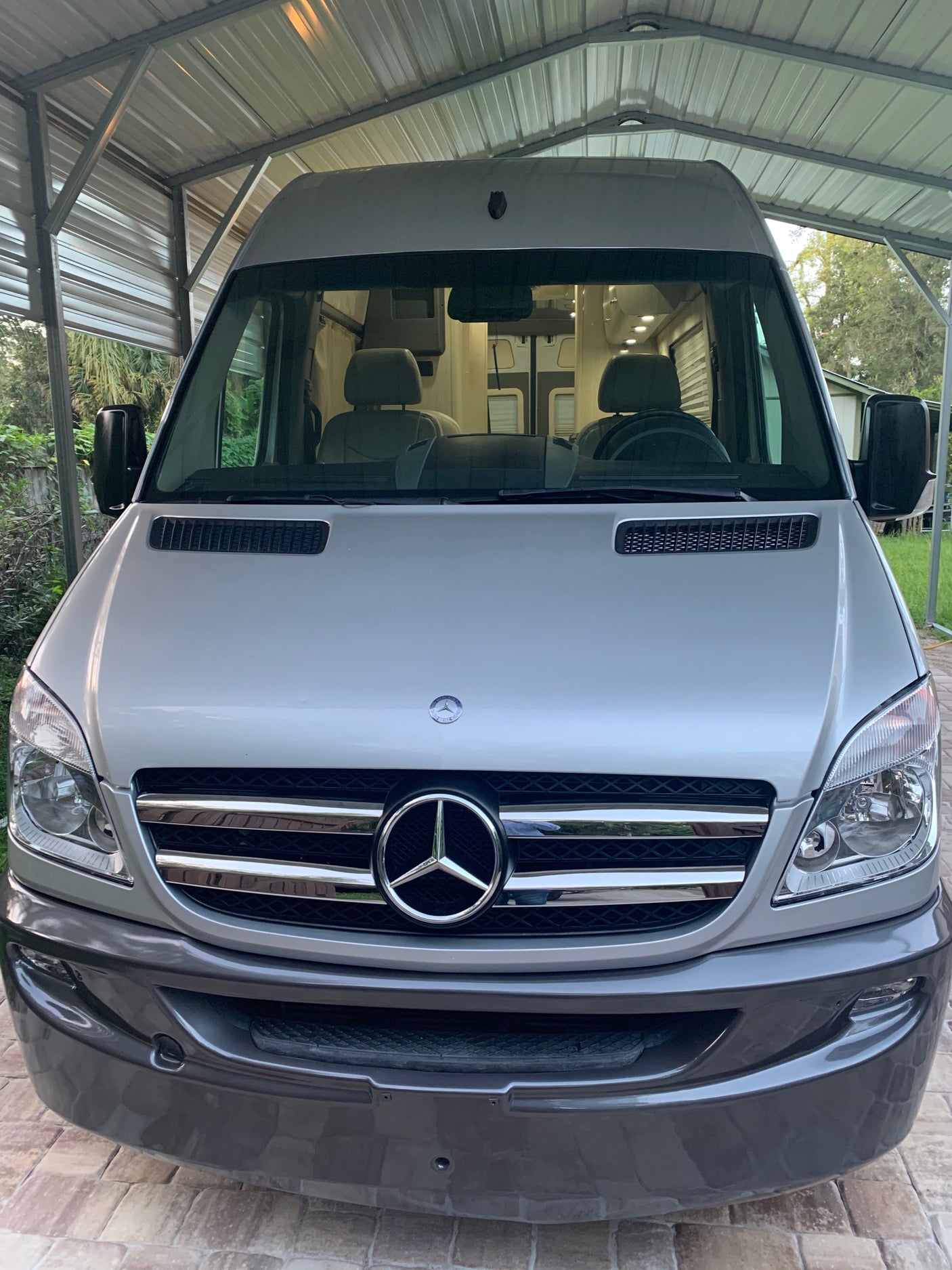 Silver Mercedes-Benz Sprinter camper van under a carport. Its front shows the large grille and headlights, ready for RV rental.