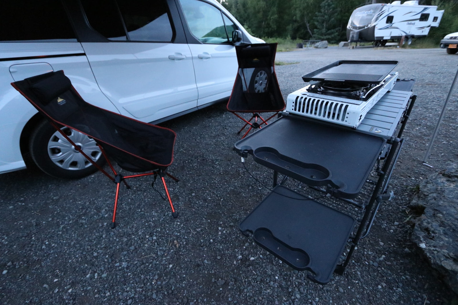An RV rental van with a compact outdoor kitchen, griddle, and two black camping chairs on gravel.