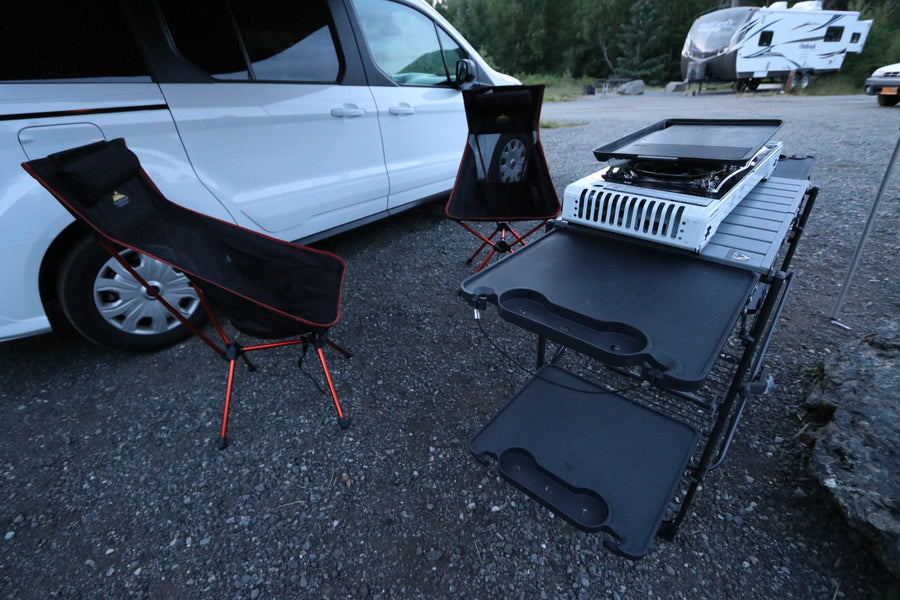 An RV rental van with a compact outdoor kitchen, griddle, and two black camping chairs on gravel.