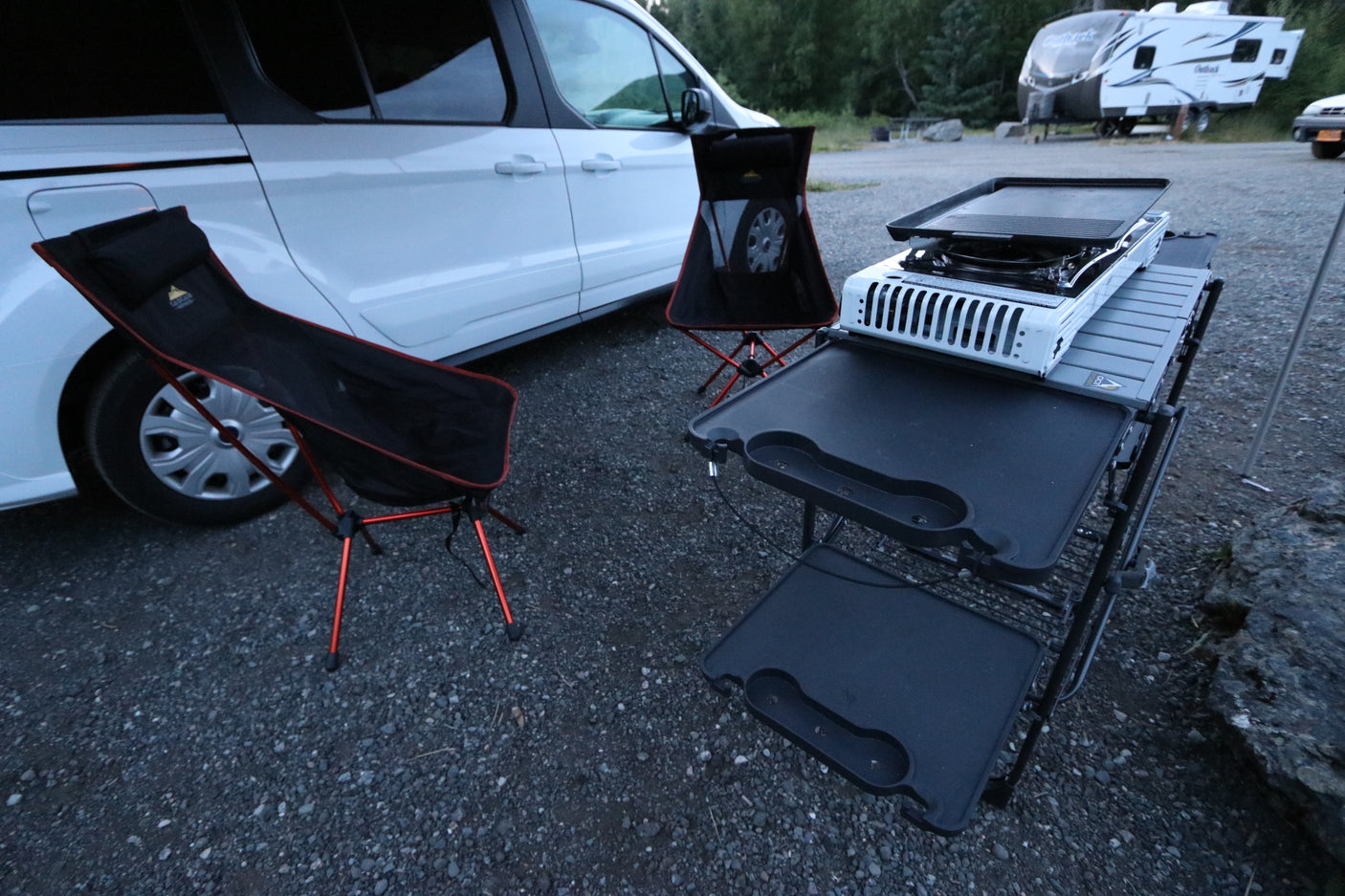 An RV rental van with a compact outdoor kitchen, griddle, and two black camping chairs on gravel.