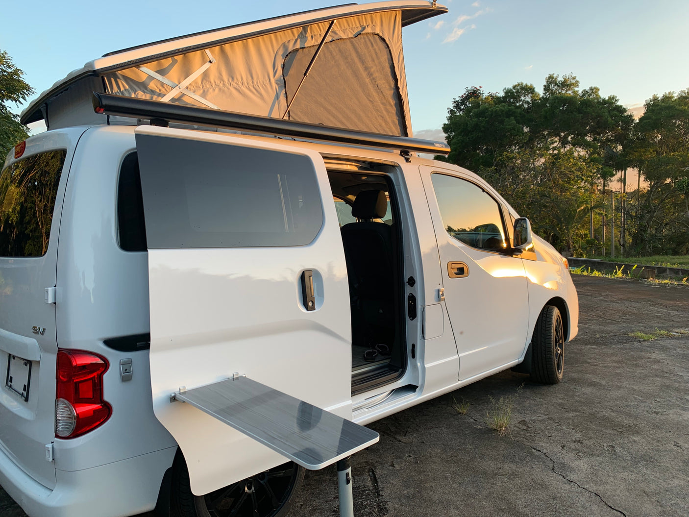 White pop-top camper van, with side door open and fold-out table, parked at sunset. Perfect RV rental for adventure.