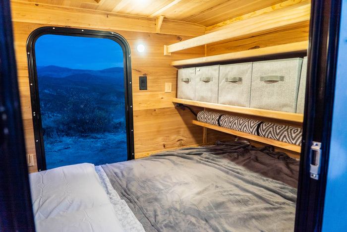 Cozy RV rental bedroom with a large window offering nature views, practical wooden shelves, and organized storage bins.