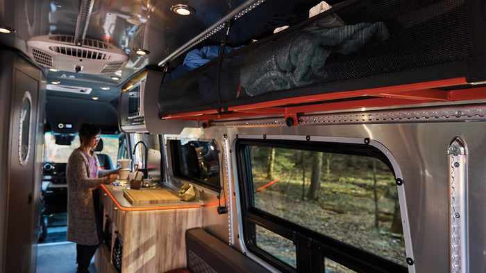 Experience an RV rental: a cozy interior with a person making coffee, forest views, and smart storage.