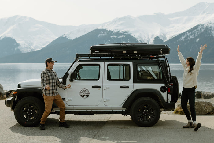 Joyful couple celebrates their Alaska RV rental adventure. White Jeep with Roofnest tent by a beautiful lake.