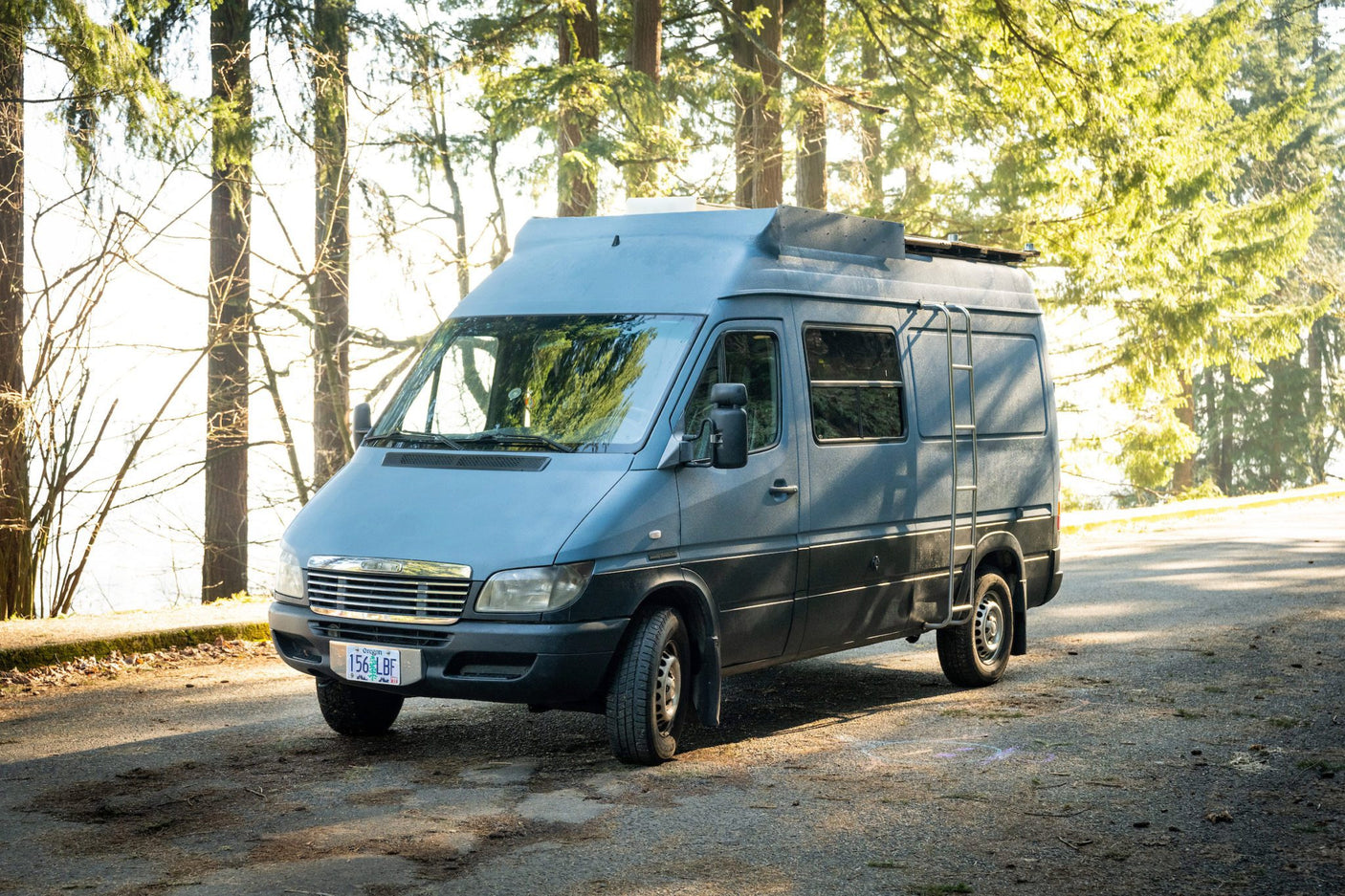 A large, custom gray Mercedes Sprinter camper van for rv rental, parked on a sunlit road in a forest. Perfect for travel adventures.