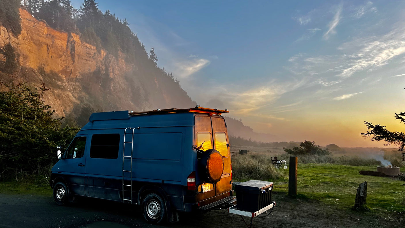 Blue RV rental parked at sunset by a vast, misty coastal cliff. A campfire glows in the grassy field nearby.