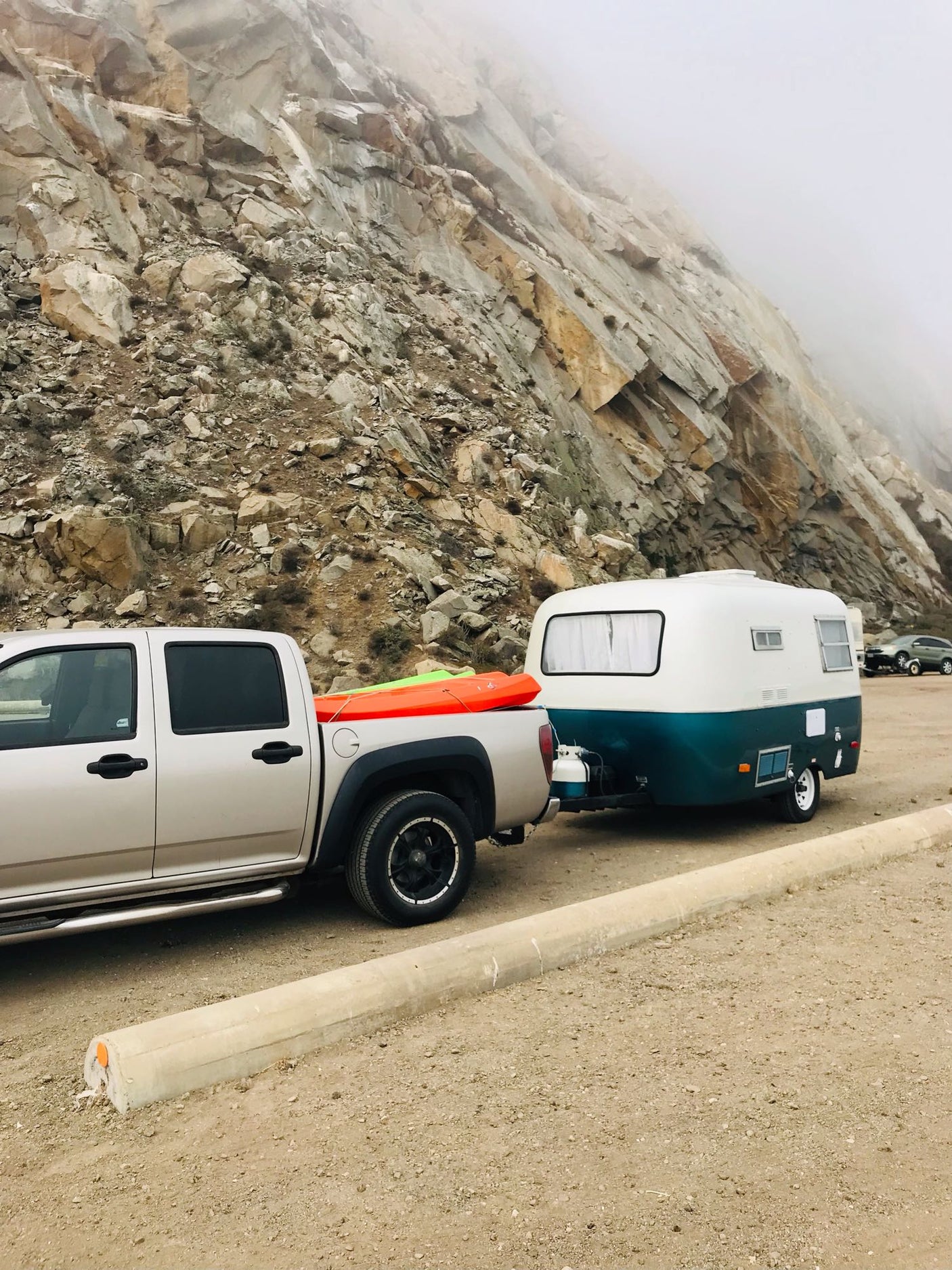 Explorer's RV rental: Truck with kayaks and compact camper trailer parked against a large, misty rocky mountain.