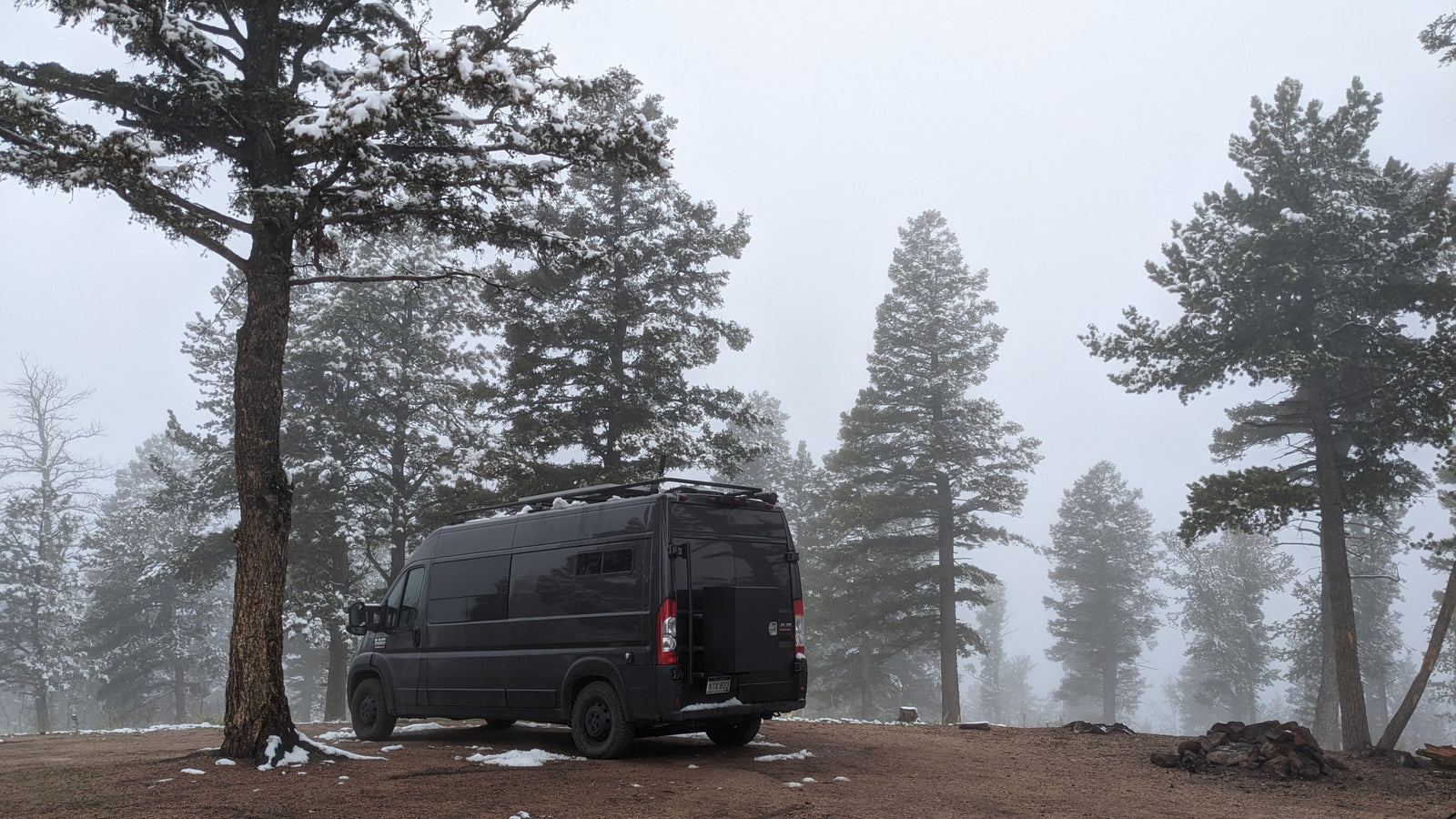 Dark camper van, ideal for rv rental, parked in a serene, snow-dusted pine forest on a foggy winter day.