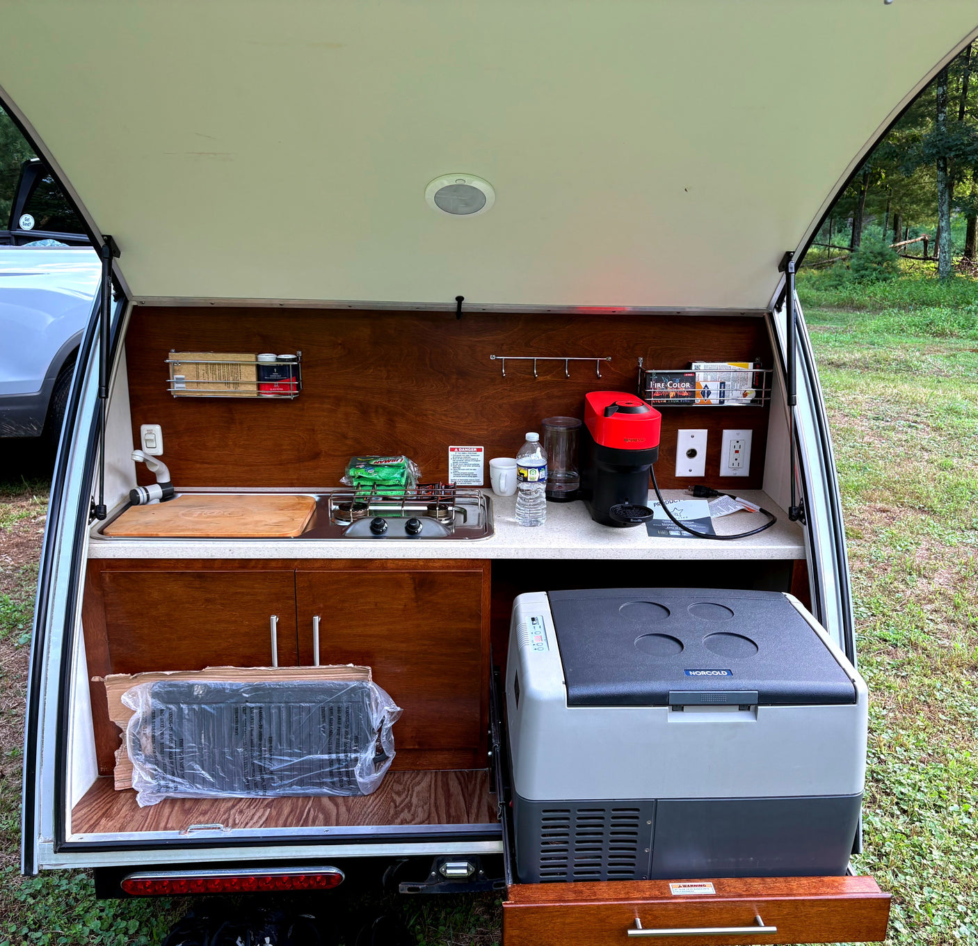 Compact RV rental teardrop trailer outdoor kitchen with Nespresso, stove, and cooler, perfect for camping.