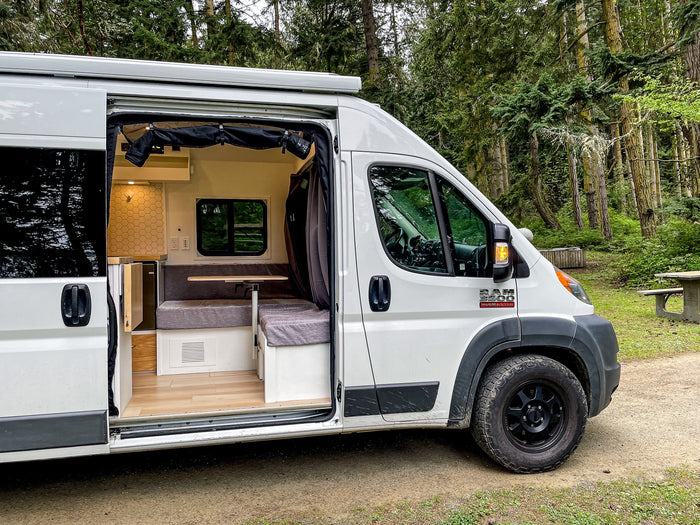 This **rv rental** camper van in a forest reveals its cozy interior, featuring a kitchen, bench seating, and wooden floor.