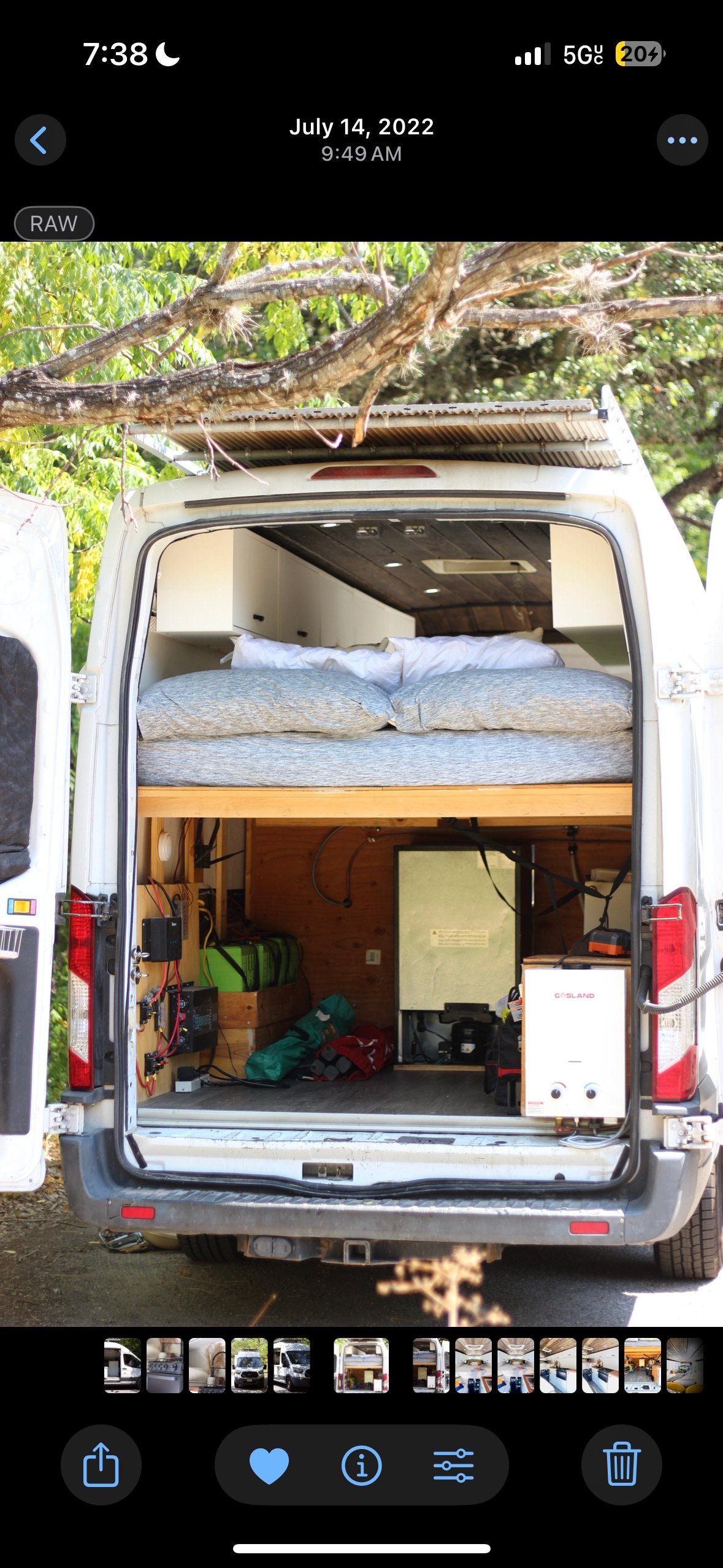 Rear view of an RV rental: a white conversion van with open doors showing an elevated bed, batteries, fridge, and water heater.