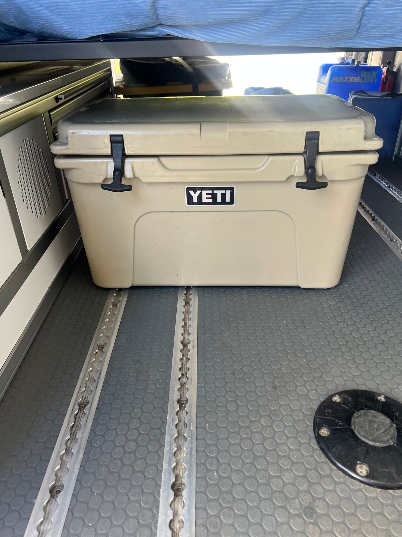 Large beige Yeti cooler inside an rv rental, resting on a textured floor with metal securing tracks. Other gear visible in the back.