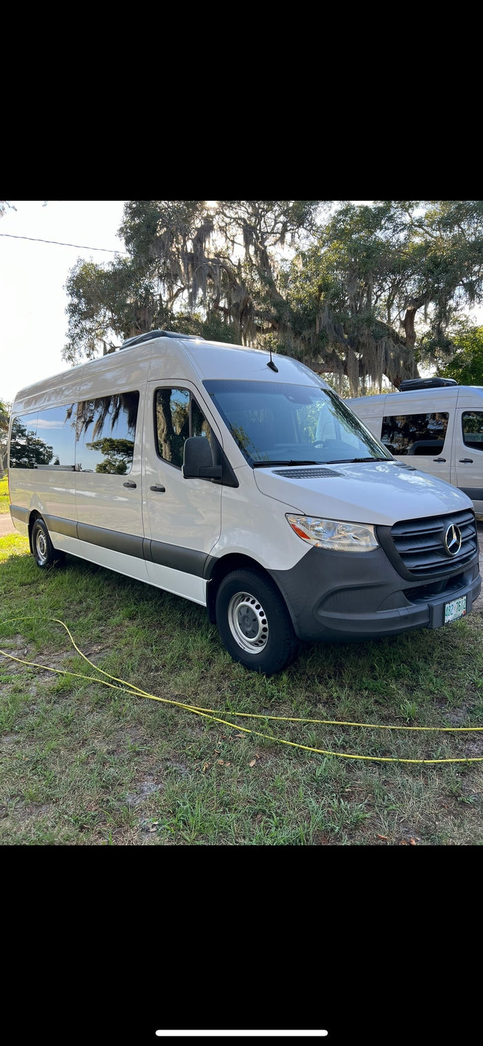 This white Mercedes Sprinter van, a modern RV rental, is parked on a grassy lot with mossy trees and another van behind.