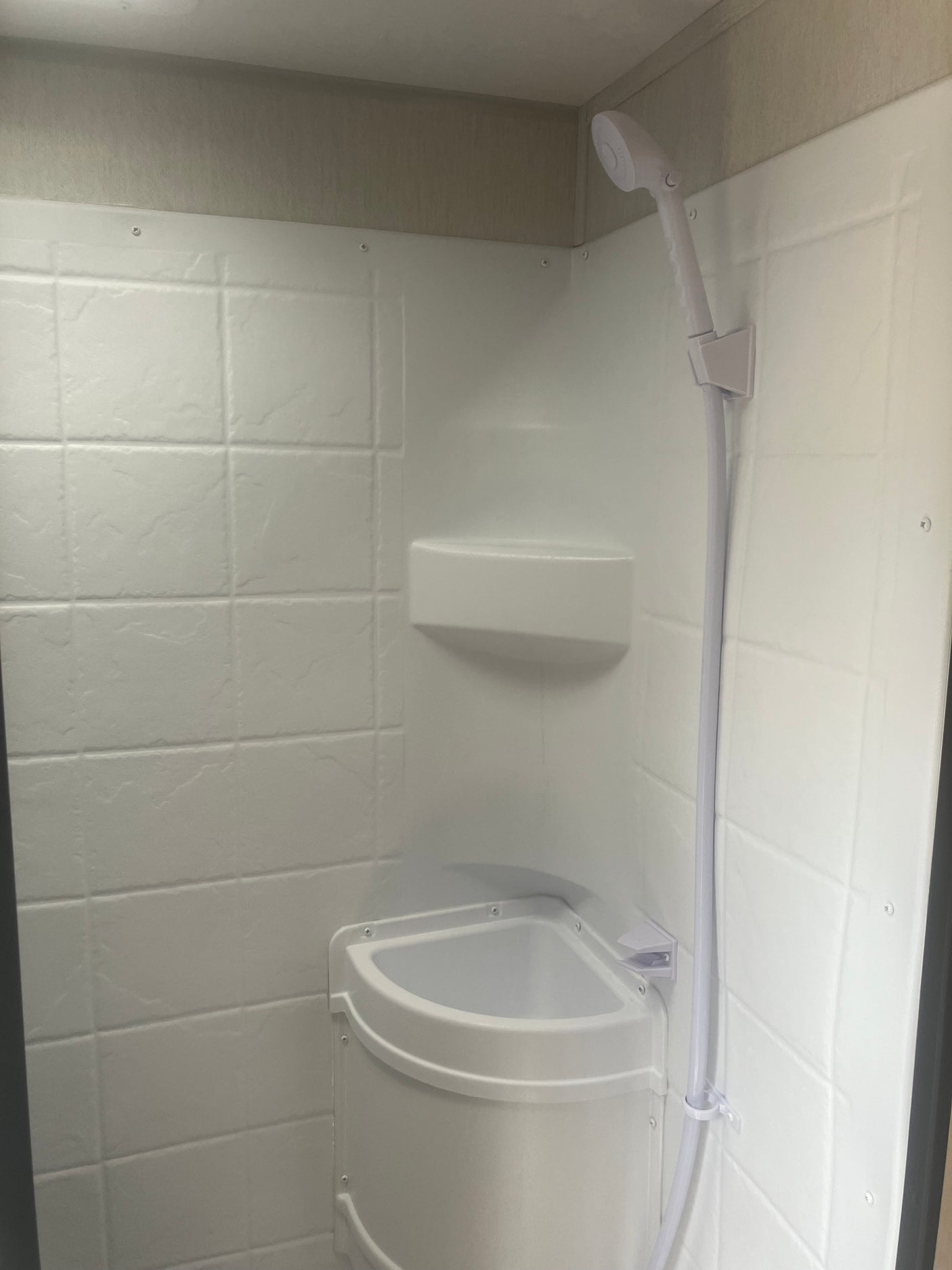 Compact white shower in an RV rental, featuring a handheld shower head, corner shelf, and textured wall panels.