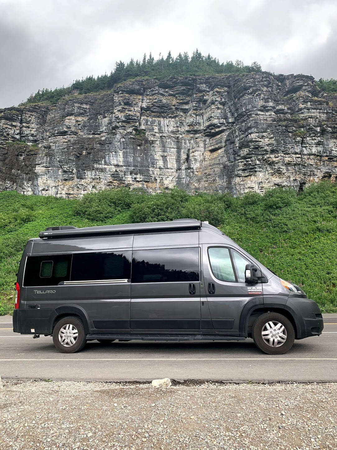 Dark grey camper van RV rental parked on a road beside a massive rocky cliff covered in green foliage and trees.