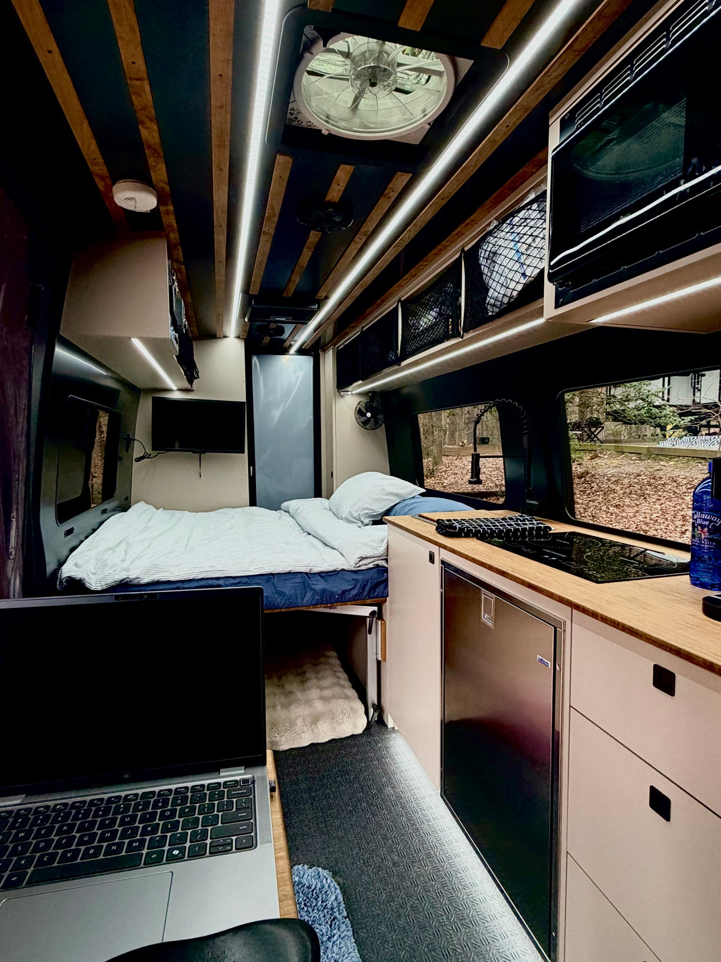Cozy RV rental interior: laptop workspace, comfy bed, kitchenette with fridge & cooktop. Ideal for travel.