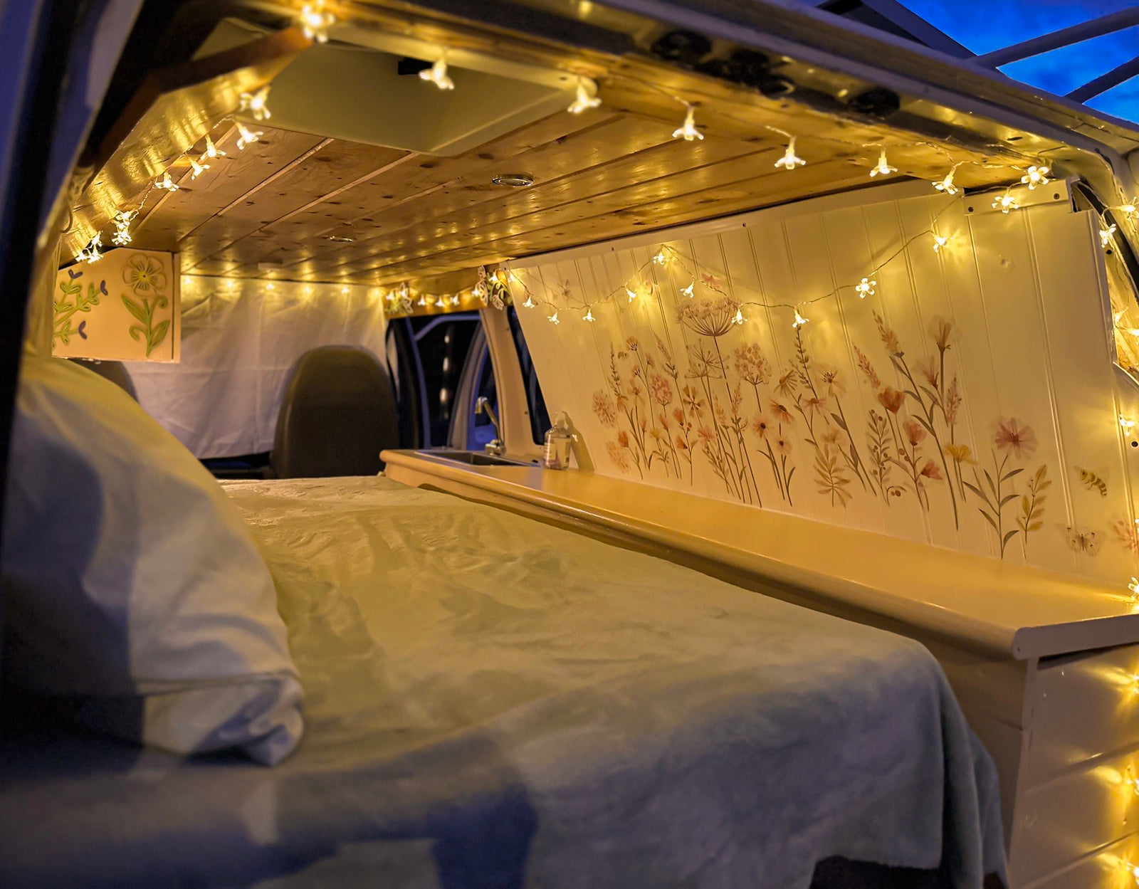 Cozy RV rental interior with flower string lights, bed, and kitchenette. Floral wall decor & wooden ceiling create a warm, inviting space.