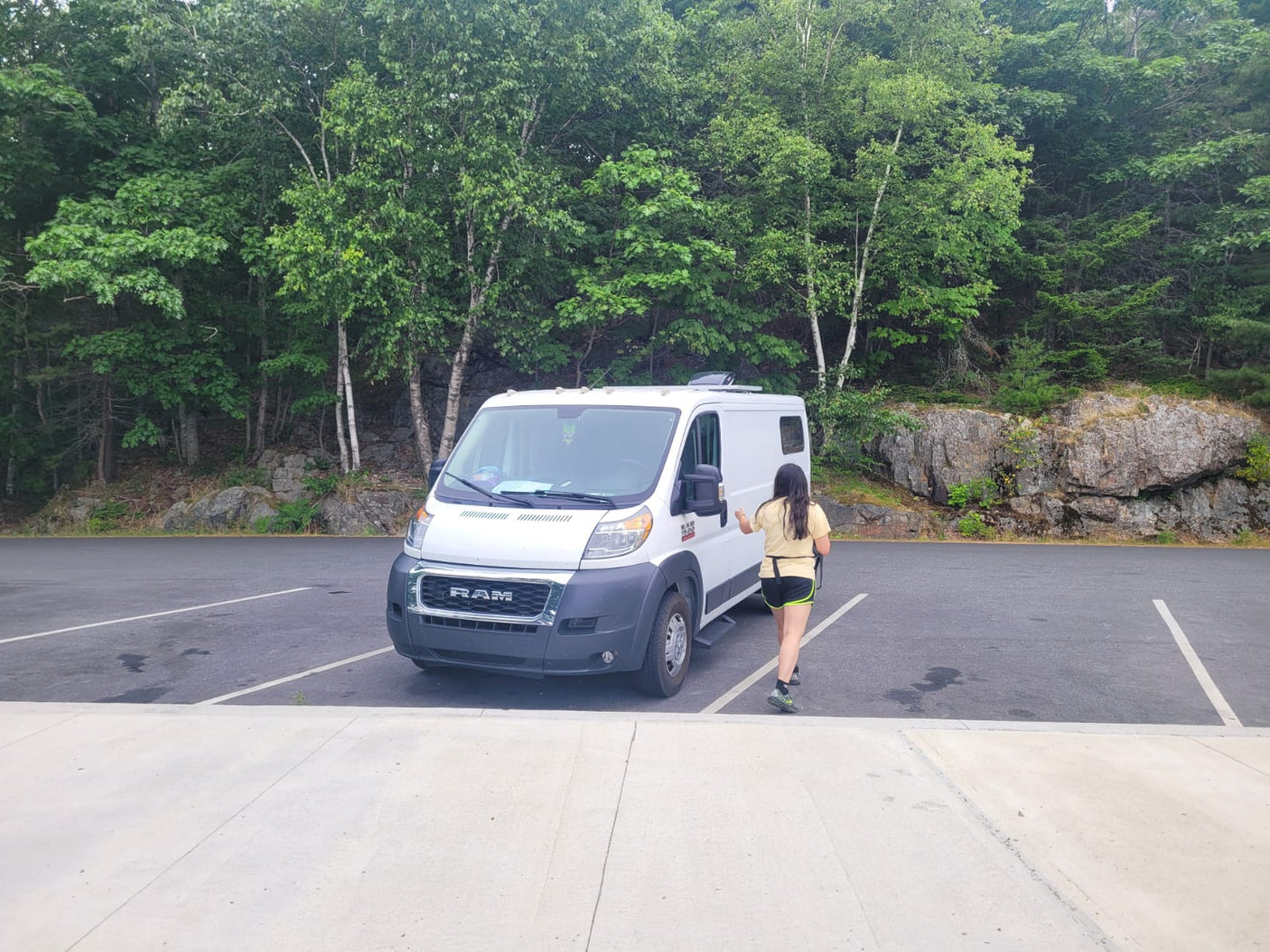 Woman approaching white RV rental van in a scenic parking lot with trees & rocks.|Cozy RV rental kitchen & sleeping nook features a modern black sink