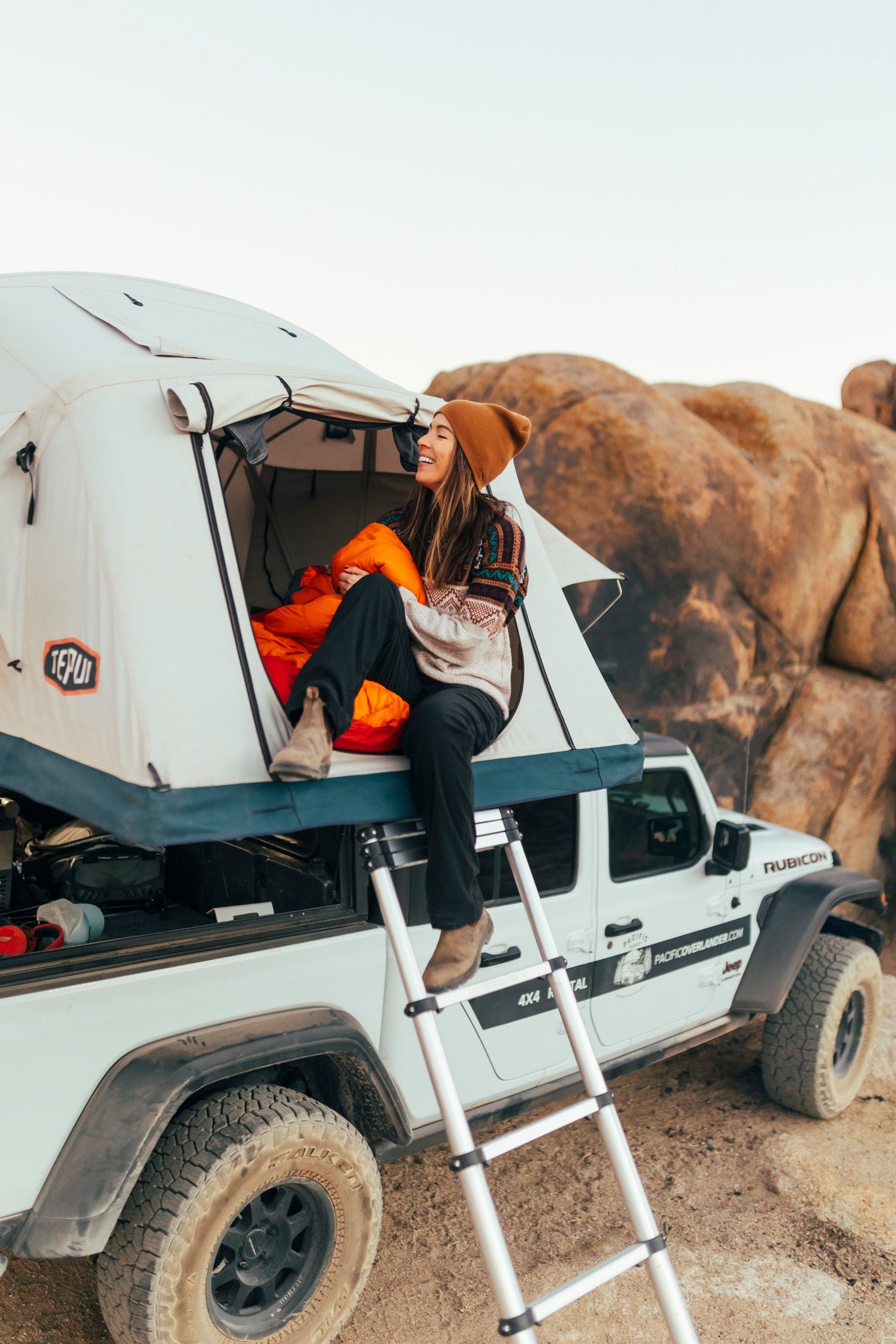 Pacific Overlander RV rental: Woman cooks from a customized Jeep Gladiator in a vast desert landscape.|Starry night camping in a 4x4 rental! Person entering a Tepui rooftop tent on a white Jeep Gladiator. Perfect for your RV rental adventure.|A cozy desert camp at dusk: white Jeep with rooftop tent and bright campfire