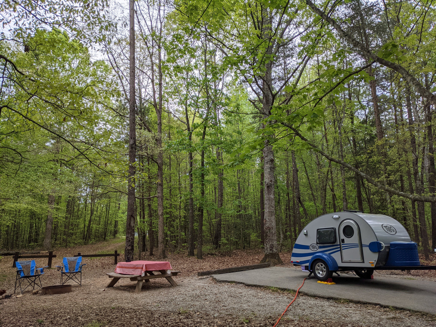 Serene campsite with a teardrop RV rental parked on a paved pad, complete with a picnic table and lush trees.