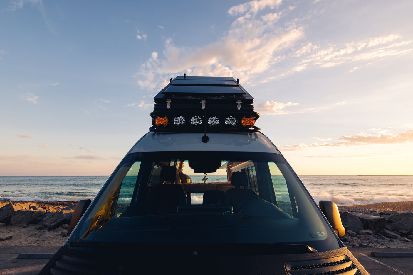 An RV rental camper van with rooftop solar panels & lights overlooks the ocean at golden hour.
